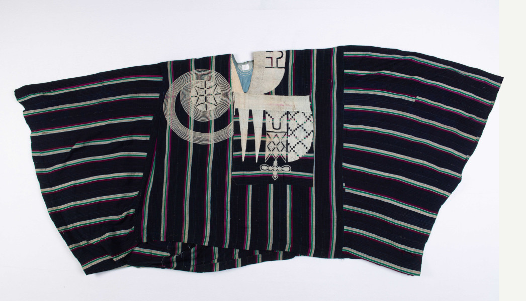 Boubou, voor 1986 gemaakt in Nigeria, van geweven indigo geverfde katoenen banden met zijde borduursels. Het naaiwerk is machinaal. De twee naar beneden wijzende punten stellen messen voor en geven aan dat de drager enig aanzien had. (WM-70058)